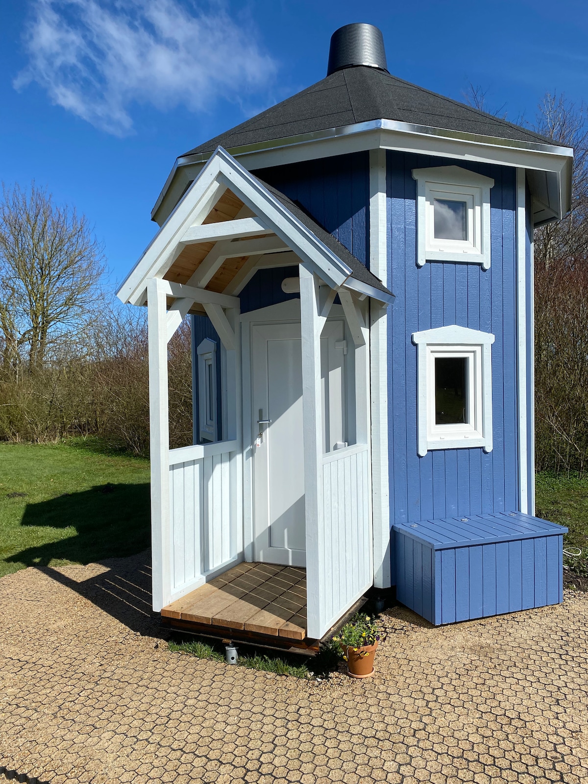 A blue tiny house features a distinctive tower shape and an inviting entry with a white door. Small rectangular windows add charm, and a wooden porch leads to the entrance. Soft grass and a potted plant are visible at the base, set against a clear blue sky.