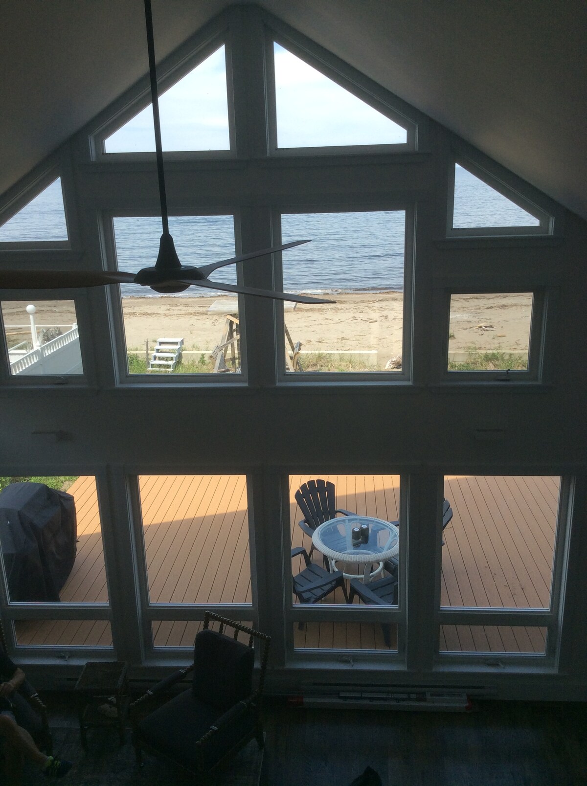 Large windows allow for natural light to fill the space, offering a view of the sandy beach and water beyond. A patio table and chairs are positioned on the deck, while a grill is visible nearby, promoting outdoor enjoyment.