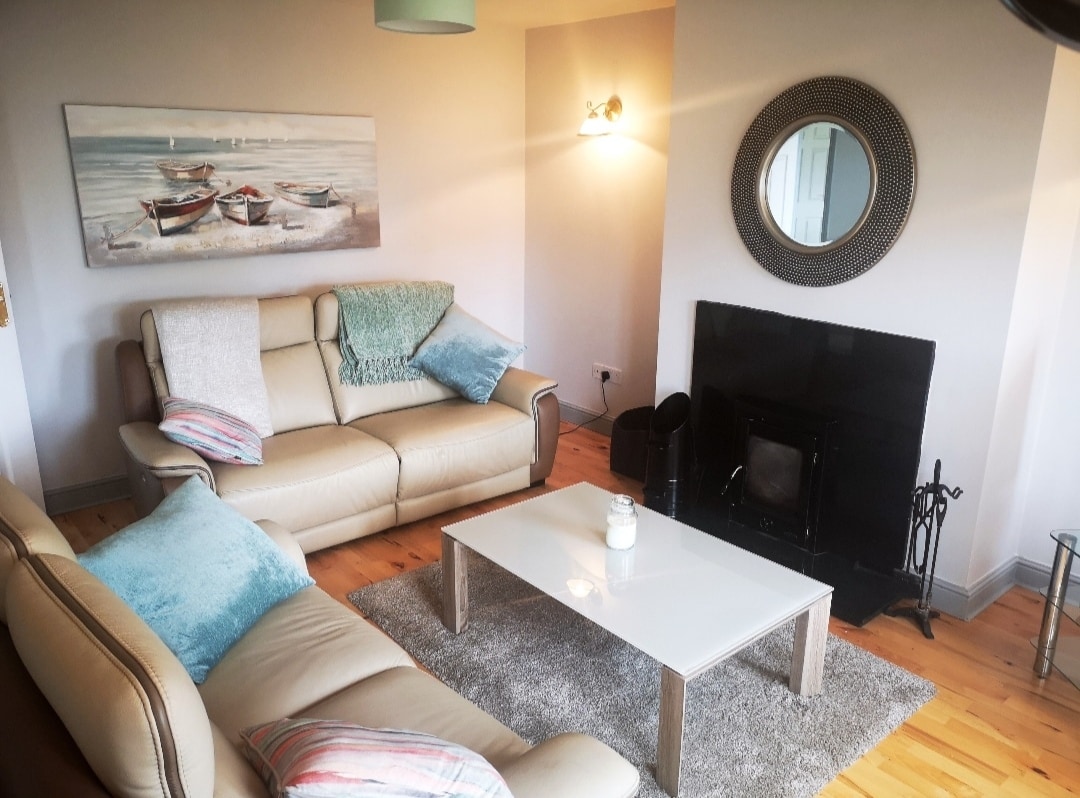 A comfortable living area features light-colored leather sofas arranged around a glass coffee table. A soft rug is placed beneath the table, and a painting of boats complements the bright space. A round mirror and a fireplace are also visible, enhancing the inviting ambiance.