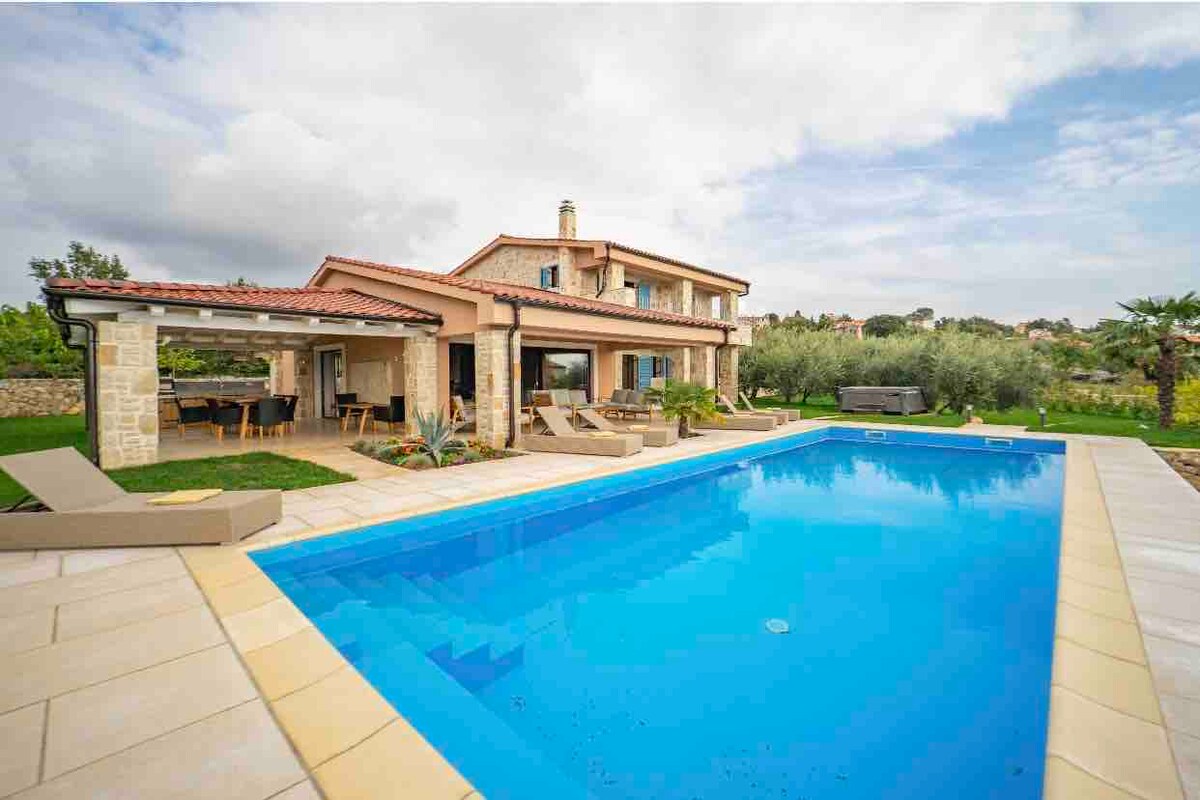 The modern stone villa features a private saltwater pool surrounded by lounge chairs. An outdoor dining area is covered by a roof and overlooks the pool. Lush greenery of an olive orchard is visible in the background, showcasing a tranquil outdoor environment.