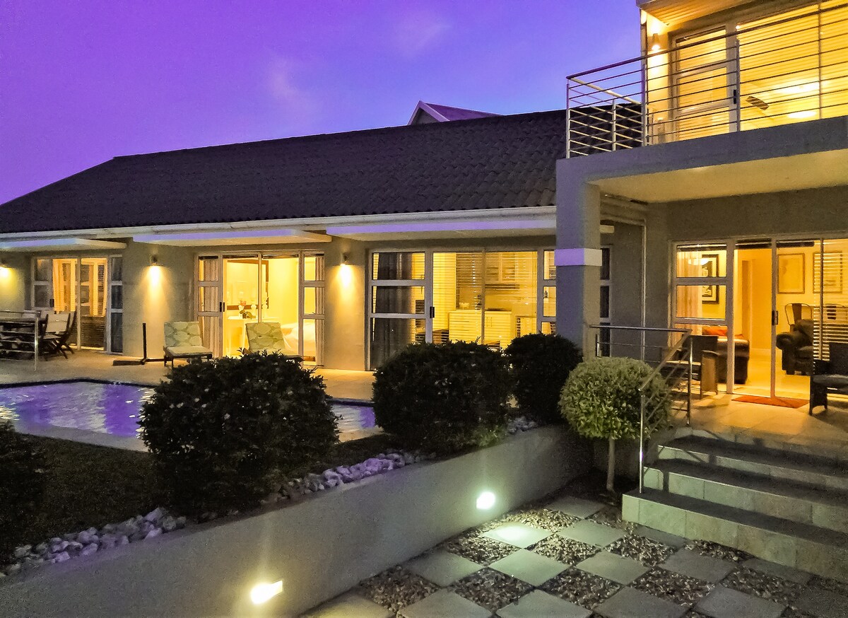 A modern villa is depicted at dusk, featuring a well-lit interior visible through large windows. A private pool is situated in the foreground, surrounded by neatly arranged landscaping. The entrance is framed by manicured shrubs and stone steps leading to the upper level.