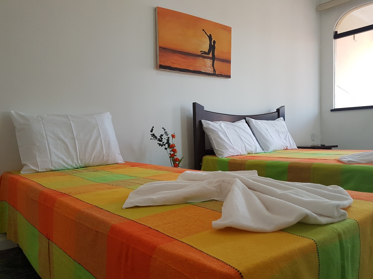 A bright sleeping area features two beds with colorful coverings in vibrant orange and green. Fresh white linens are neatly arranged on each bed. A piece of wall art depicting a sunset scene and a dancer is displayed above. A small flower vase adds a touch of freshness.
