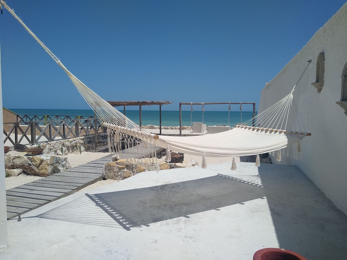 A spacious outdoor area features a large hammock suspended between two posts, overlooking the ocean. A wooden deck leads towards a beachside lounge space, which includes a shaded seating area under a rustic pergola. Bright blue sky complements the tranquil coastal setting.