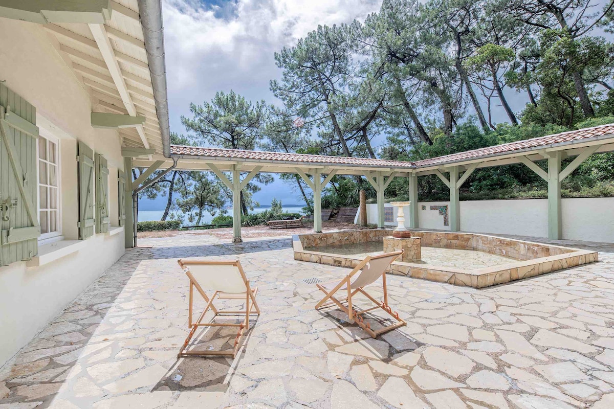 A spacious outdoor area is paved with natural stone, featuring a central stone basin. Two wooden lounge chairs are positioned invitingly under a covered terrace. Surrounding trees provide a backdrop of greenery, with views of the distant landscape framed by the structure's eaves.