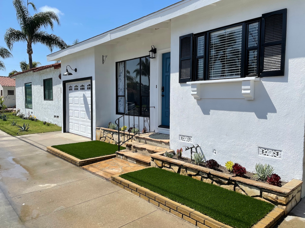 The exterior features a modern, white facade with black shutters and a bright blue door. Lush, green artificial grass outlines a stone pathway leading to the entrance, complemented by low-maintenance succulent gardens on either side. A garage is visible to the left.