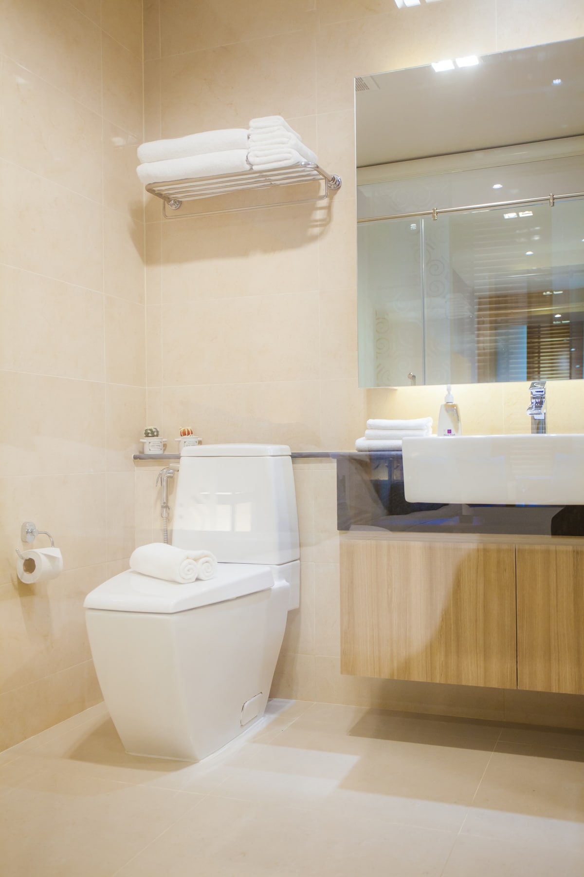 A modern bathroom features neutral-toned tiles and wood accents. A white toilet is positioned next to a wooden vanity, which holds a spacious sink. Several rolled towels are neatly arranged on a shelf above, with a large mirror reflecting the balanced layout.