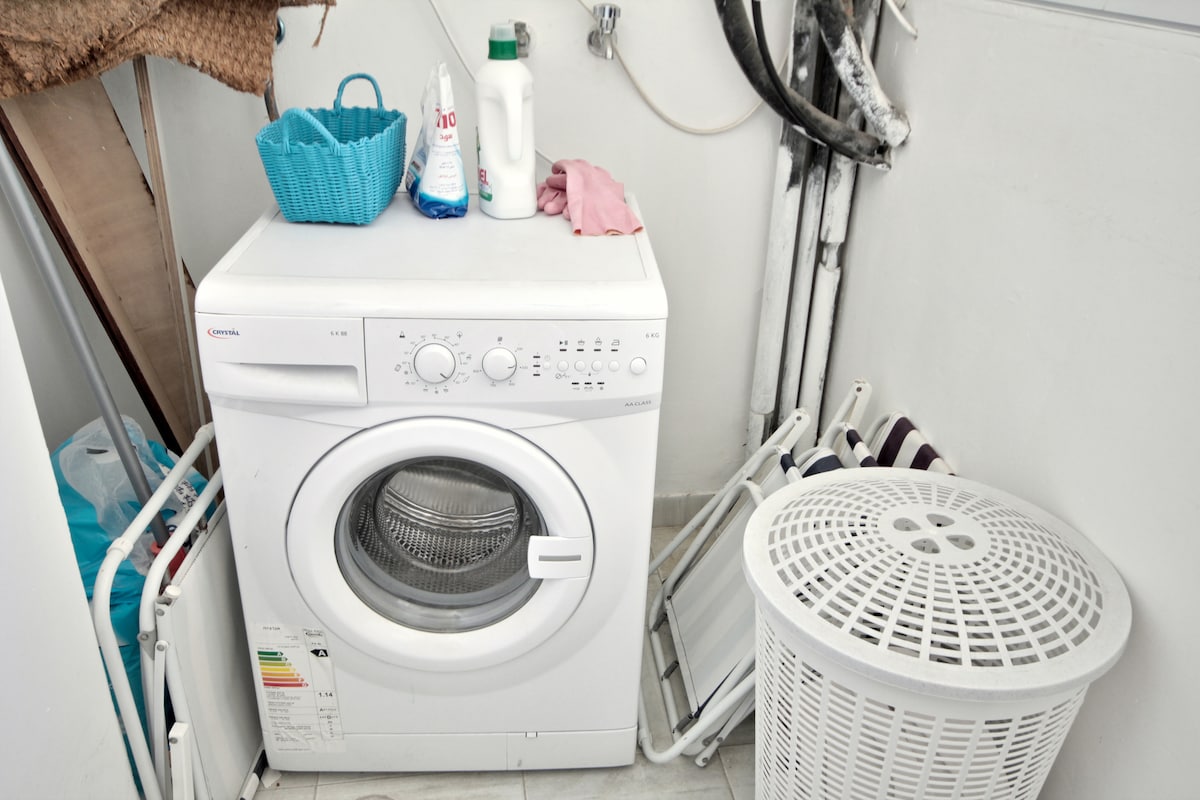 A compact laundry area is featured, showcasing a white washing machine with controls on the front. Adjacent to it, a woven laundry basket and cleaning supplies are visible, providing an organized space for household tasks. The walls are tiled in a neutral color.