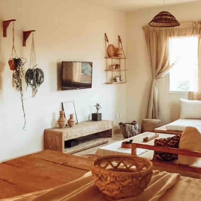 A bright living area features a minimalist design with a wooden coffee table and comfortable seating. A wall-mounted TV is positioned across from the seating, while natural light streams through a window adorned with light curtains. Potted plants and decorative items offer a touch of greenery.