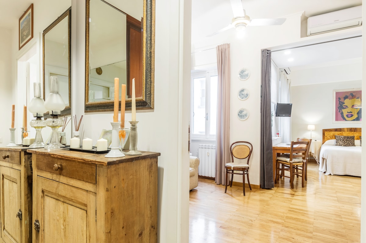 An elegant entrance area features a wooden sideboard adorned with decorative candles and vases. Beyond, a spacious living area is visible with a comfortable bed, a TV mounted on the wall, and a small dining table accompanied by a chair.