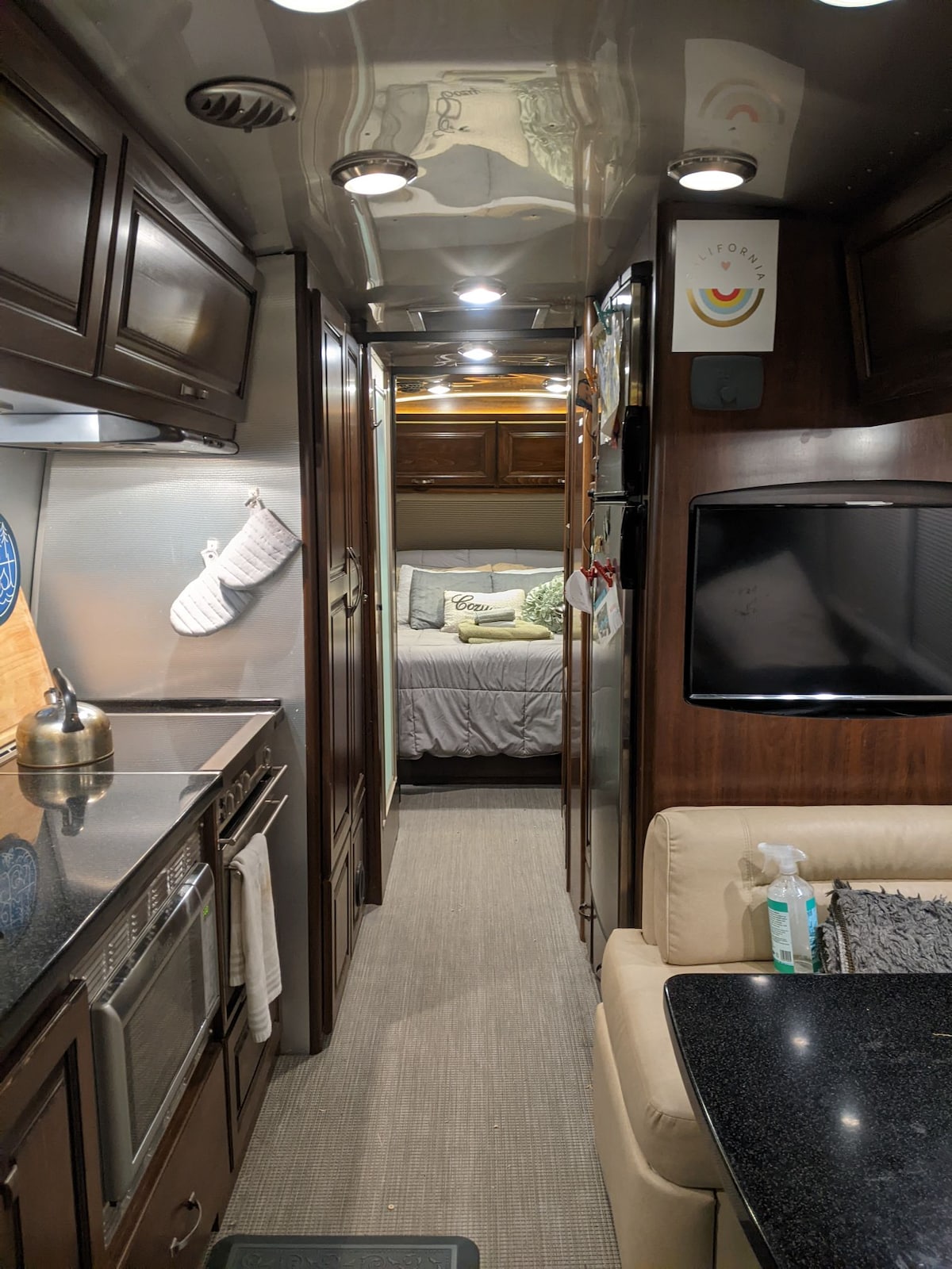 A well-designed interior showcases a narrow hallway leading to a cozy bedroom area. On one side, a full kitchen features stainless steel appliances and dark cabinetry. Opposite, a comfortable seating area with a black table is visible, creating a warm, functional living space.