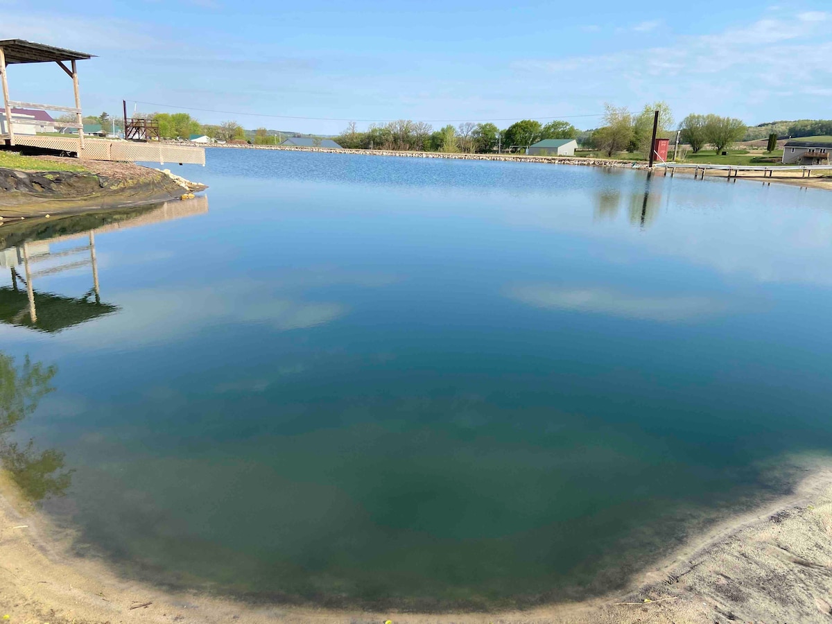 A calm pond is depicted, with clear blue water reflecting the surrounding landscape. Edges of soft sand lead to the water, and a gentle slope towards grassy areas can be seen. The backdrop includes trees and distant structures under a clear sky.