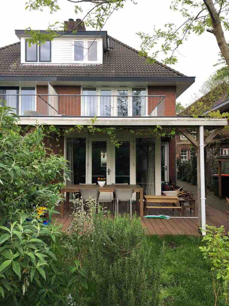 A spacious outdoor decking area can be seen, equipped with a dining table and chairs, surrounded by lush greenery. The back of the house features large windows and a balcony, allowing natural light to fill the interior spaces.
