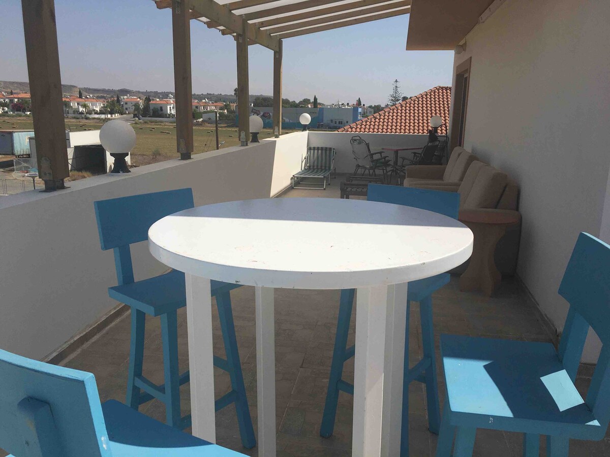 A rooftop patio is displayed, featuring a round white table surrounded by four blue chairs. A comfortable seating area with a beige sofa is positioned against the wall, while scenic views of the surrounding area are visible in the background.