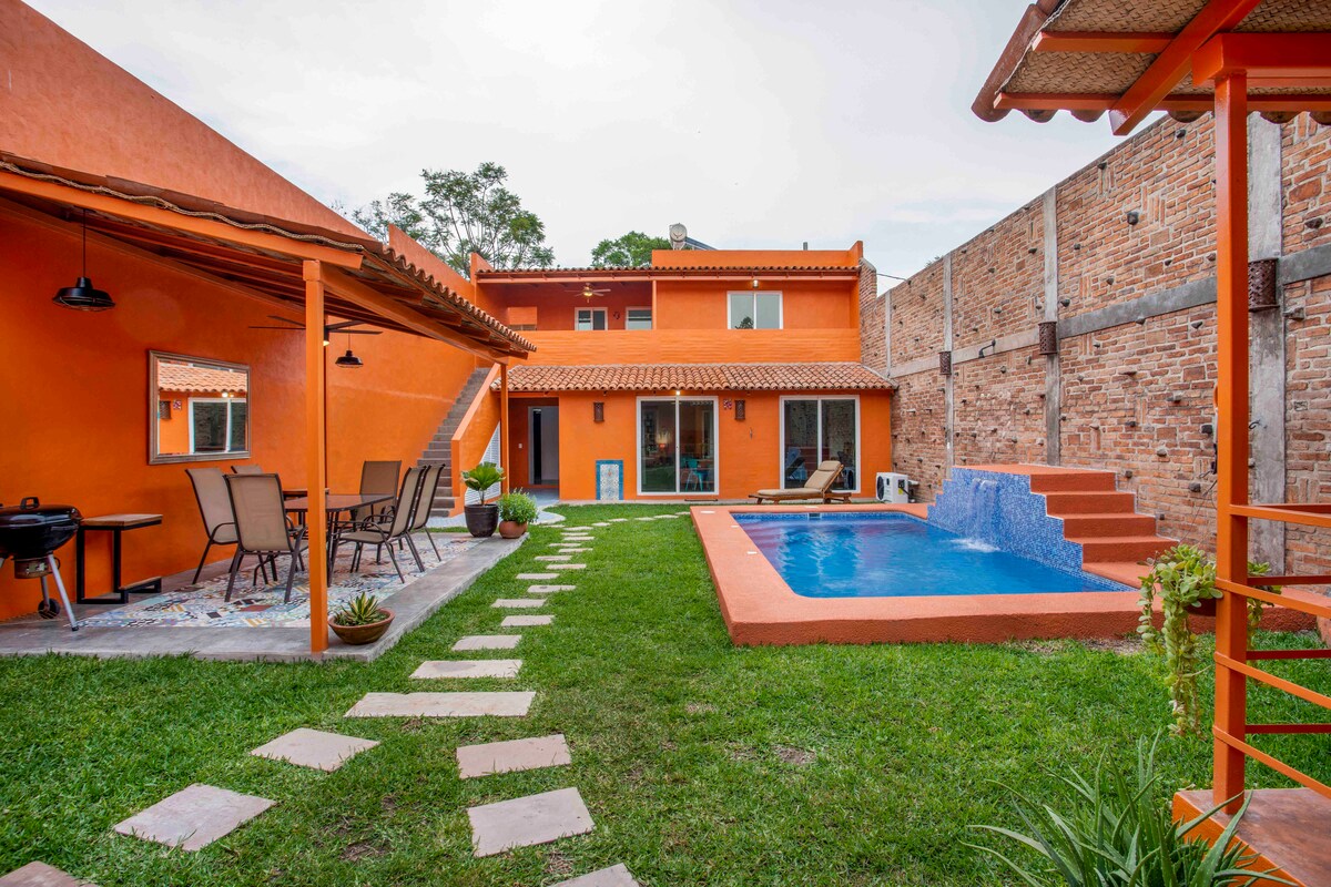 A central garden area is surrounded by vibrant orange structures, featuring a shared pool and patio. The well-maintained lawn is complemented by pathways and potted plants. Outdoor seating and a grill are positioned on the terrace, inviting relaxation in a tranquil setting.