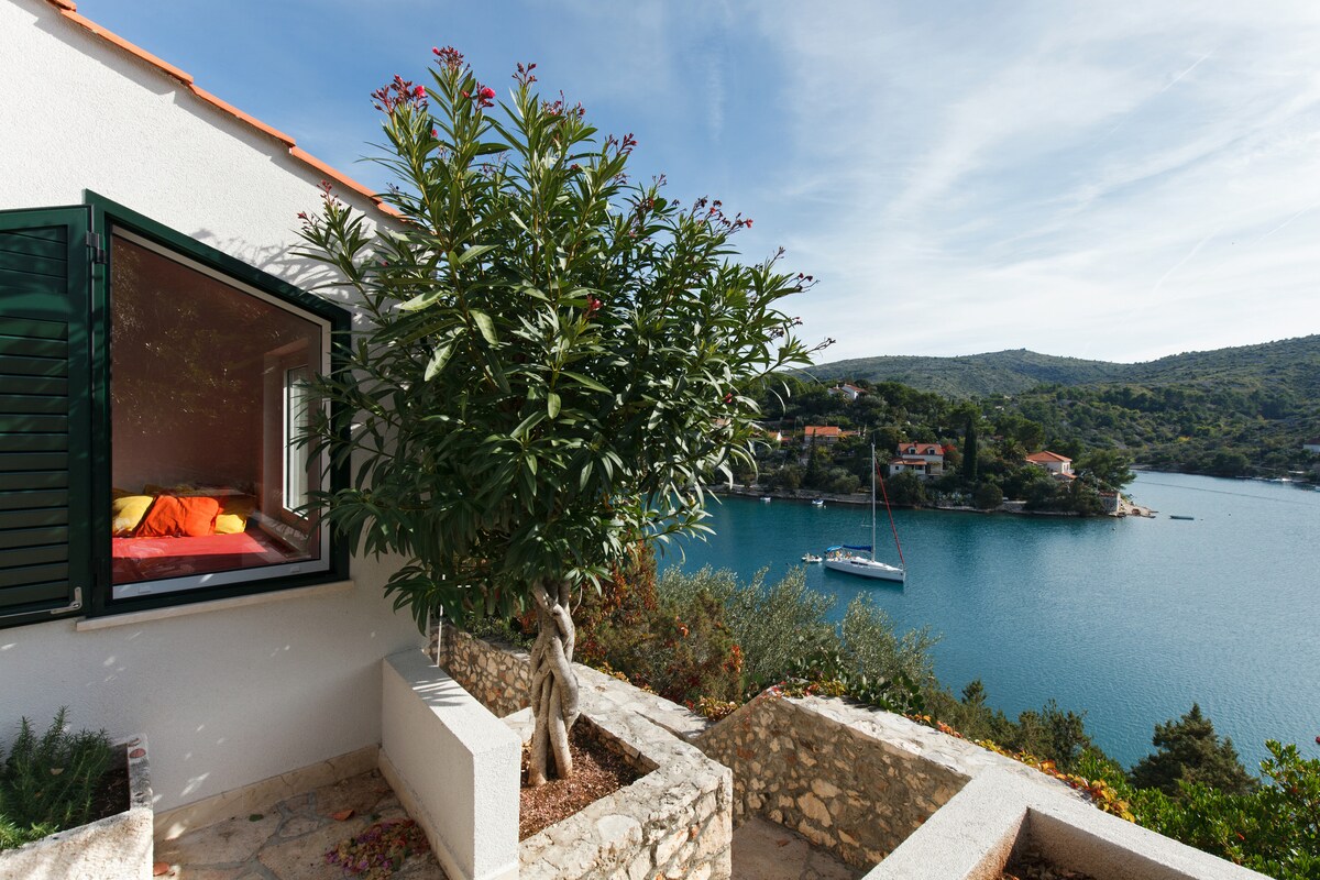 A view from the open window showcases a tranquil bay with calm waters and a sailboat anchored nearby. Lush greenery, including a flowering plant, frames the window, providing a sense of nature and serenity that complements the scenic coastline.