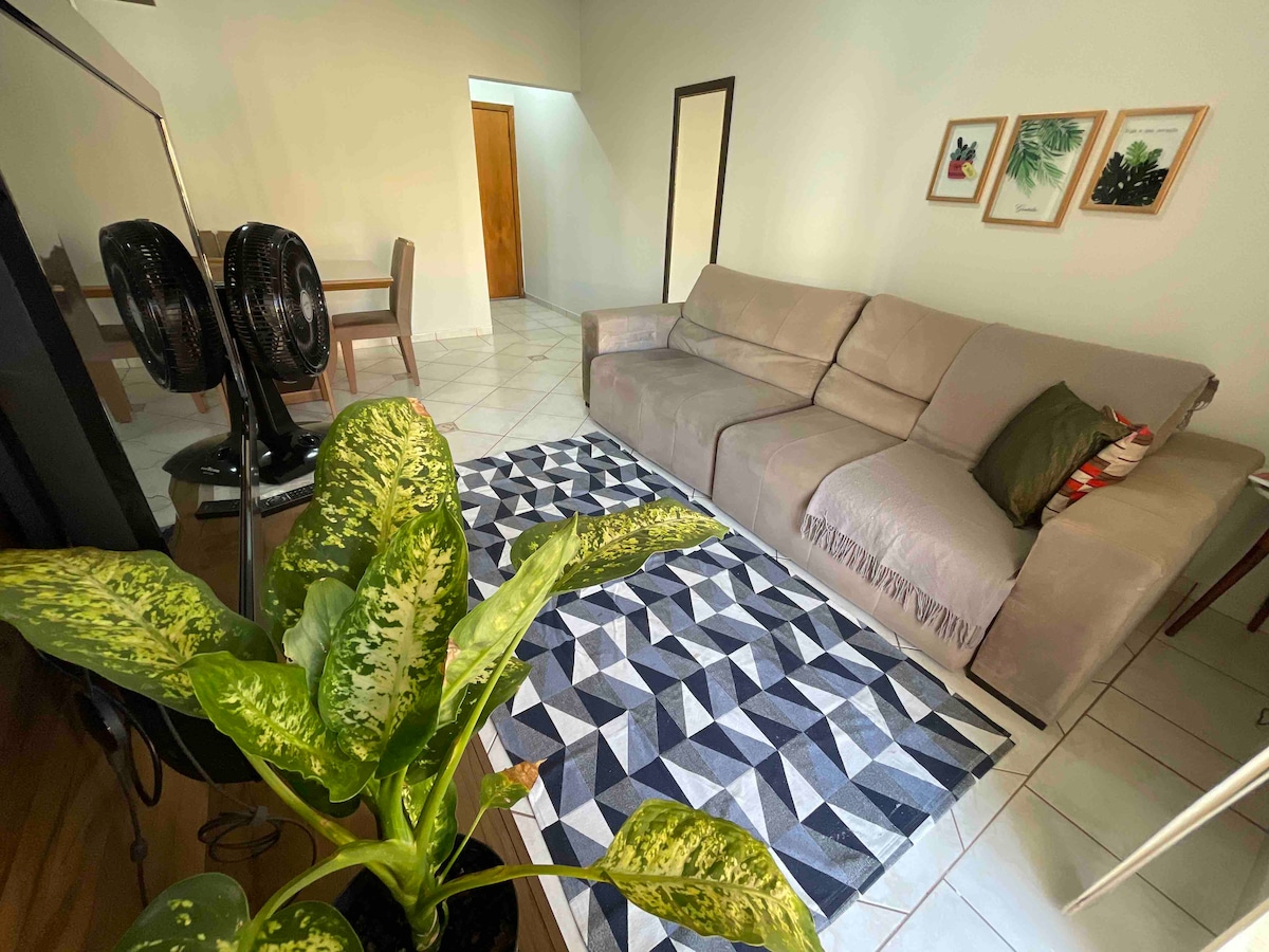 The living area features a large, comfortable sofa positioned adjacent to a patterned area rug. A small dining table is visible in the background. Two standing fans provide ventilation, and framed artwork decorates the walls. Natural light illuminates the space.
