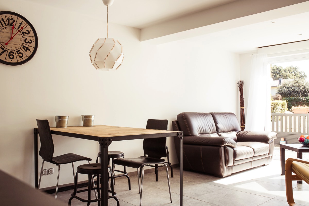 A dining area is visible with a wooden table and four black chairs, complemented by two decorative pots. A cozy leather sofa is positioned nearby, while a wall clock adds a functional element. Natural light illuminates the space through a large window.