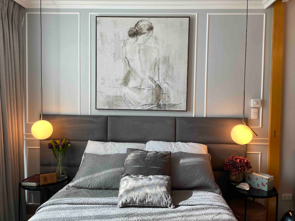 A cozy sleeping area is illuminated by pendant lights on either side of the bed. A large piece of artwork depicting a figure in a seated position hangs above a plush headboard. Decorative pillows and a small vase of tulips add subtle touches of charm.