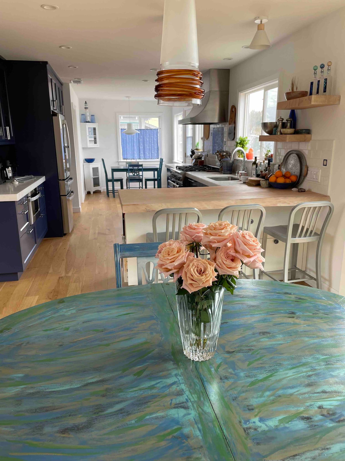 A newly renovated kitchen is showcased with modern amenities and a light color palette. A table featuring a blue-green surface is decorated with a vase of pink roses. Light streams in from large windows, illuminating the space that includes additional seating and countertops.