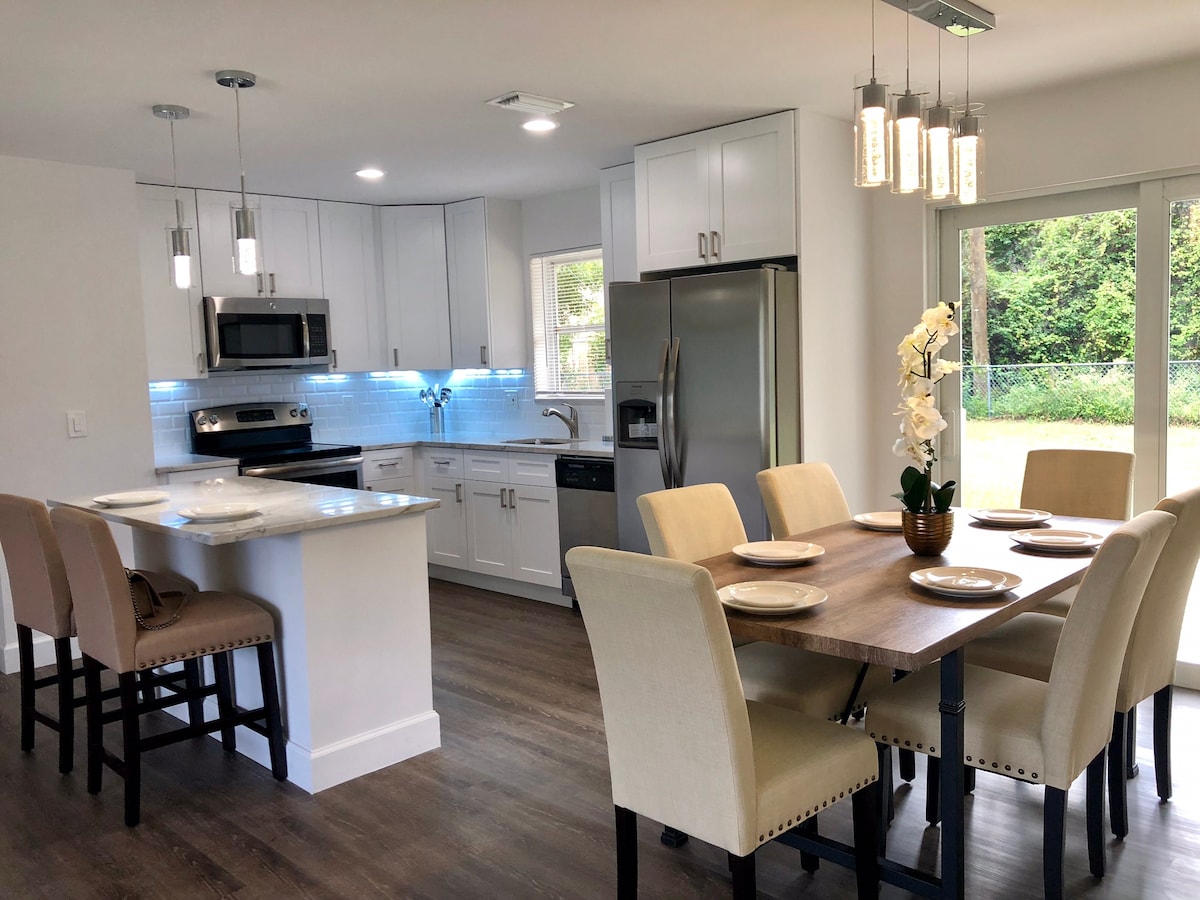 A modern kitchen and dining area is displayed, featuring sleek white cabinetry and stainless steel appliances. A large wooden dining table is set with plates, surrounded by upholstered chairs. Natural light enters through glass doors, highlighting a serene backyard view.