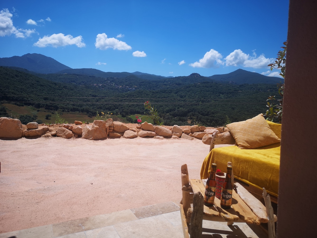 A private outdoor terrace offers a scenic view of surrounding mountains and valleys under a clear blue sky. Natural stone walls frame the space, while a rustic wooden chair is positioned beside a soft yellow throw. The area is bathed in natural light.