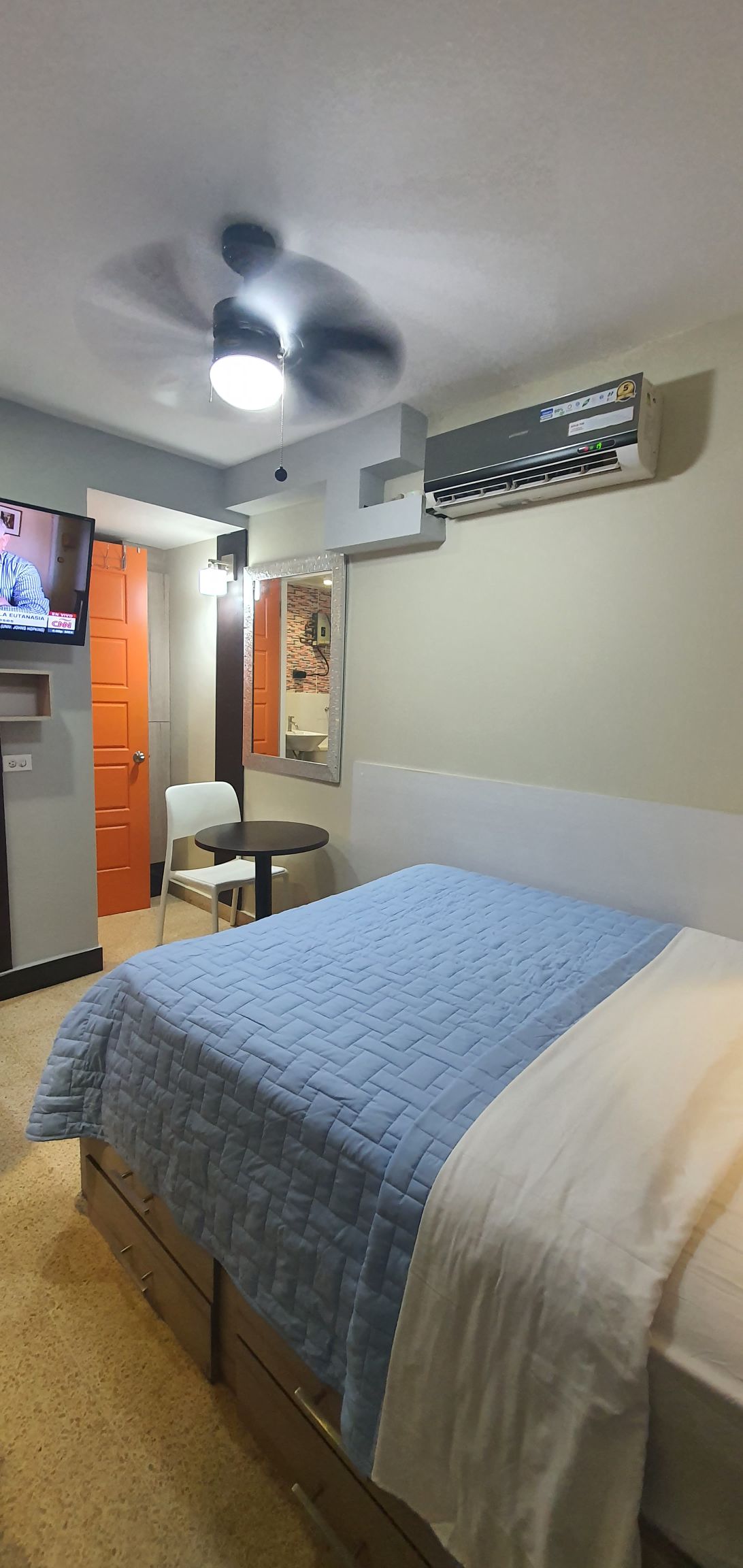 A modern studio features a comfortable bed covered with a light blue quilt. A small round table and chair are positioned nearby, while a wall-mounted television provides entertainment. An air conditioning unit is visible, alongside an orange door leading to a private bath area.
