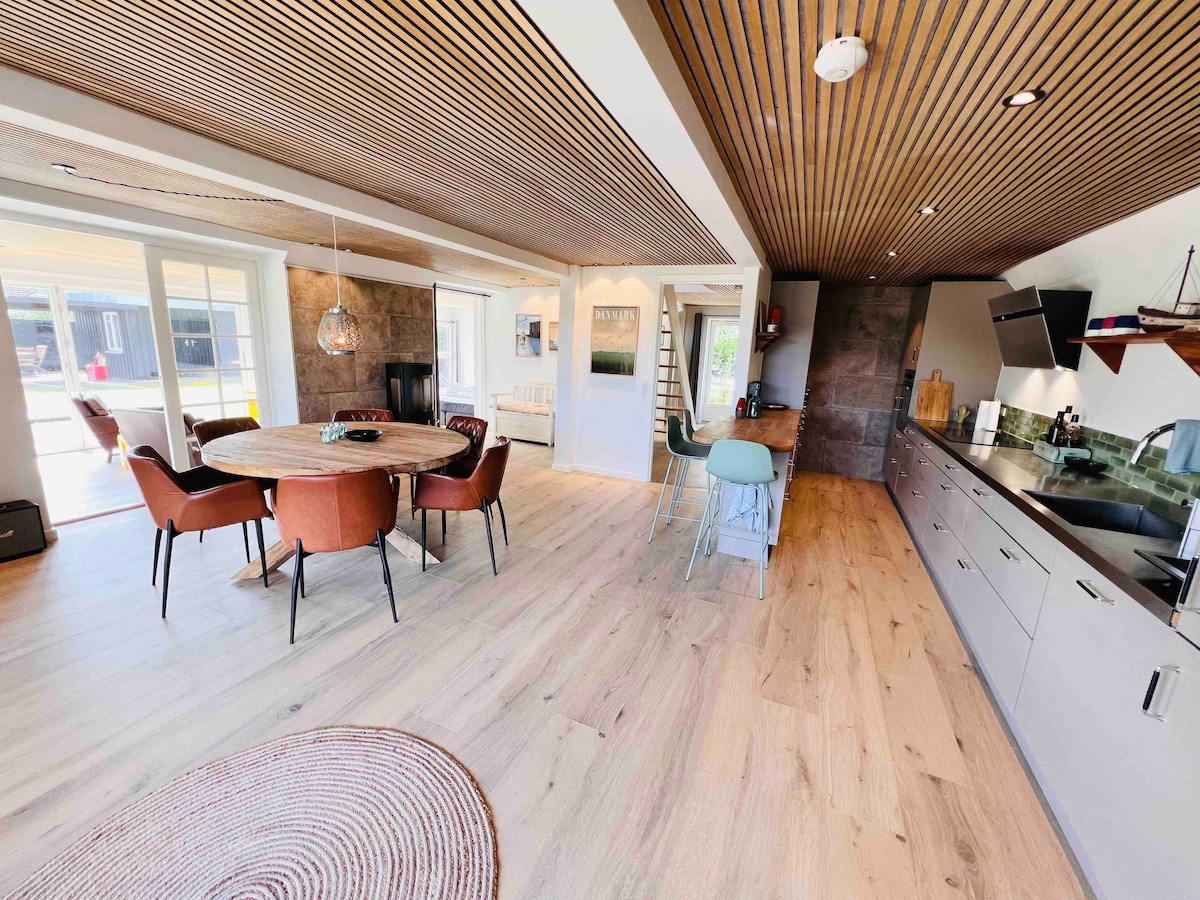 An open-concept living area features a circular dining table surrounded by upholstered chairs. A modern kitchen showcases sleek cabinetry and stainless steel appliances. Natural light floods the space through large sliding doors, highlighting the wooden ceiling and warm-toned flooring.