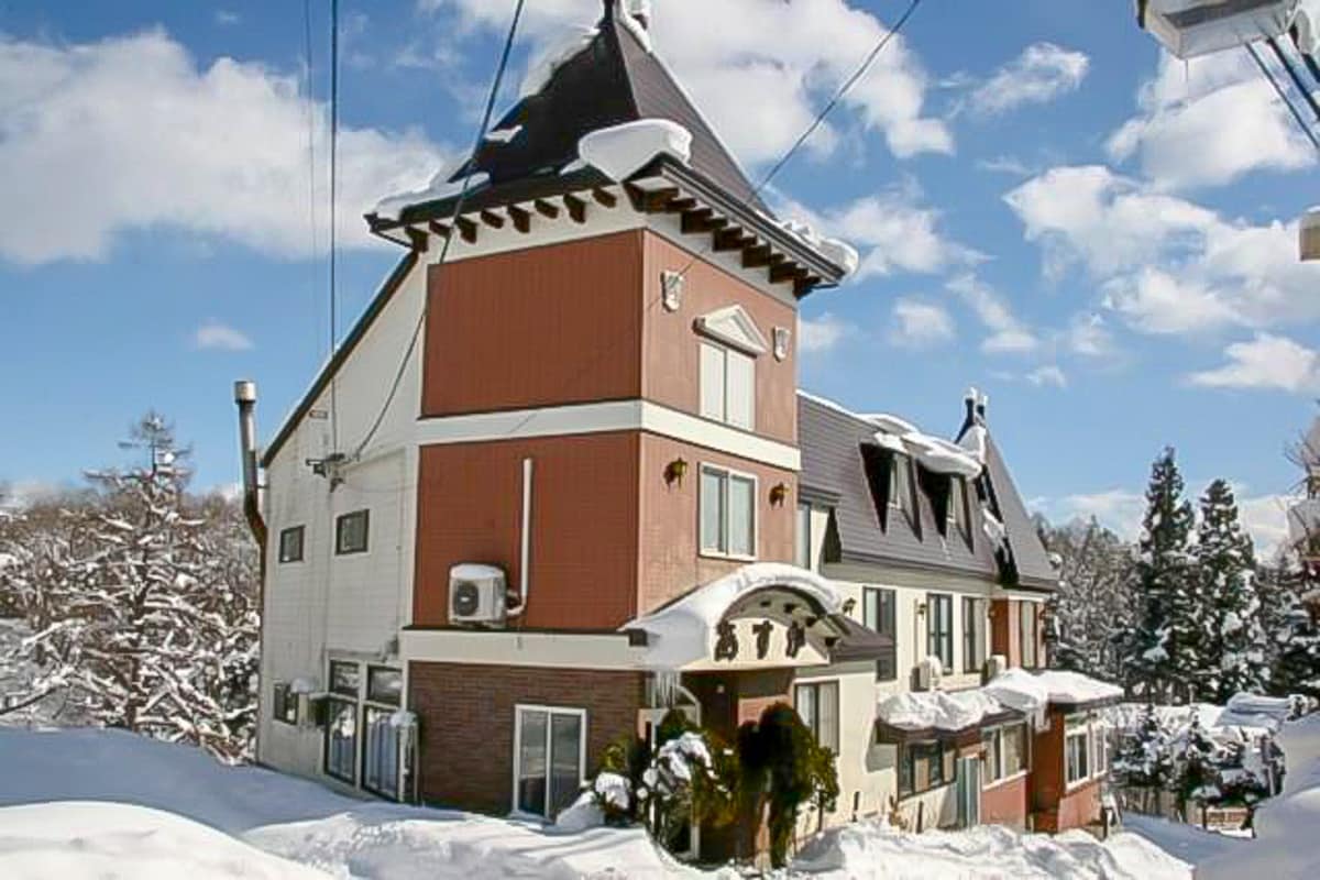 The exterior of a multi-story lodge is displayed under a clear blue sky, featuring a distinct peaked roof. Snow blankets the surrounding area, highlighting the building's warm tones and large windows. Tall trees in the background are adorned with fresh snow.