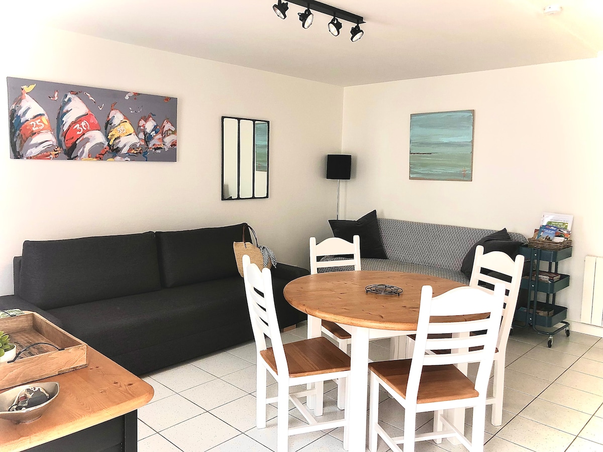 A functional living area is presented with a dark sectional sofa and a light wooden dining table surrounded by four chairs. Artistic wall decorations add a touch of color, while a standing lamp and a corner armchair provide additional seating options.