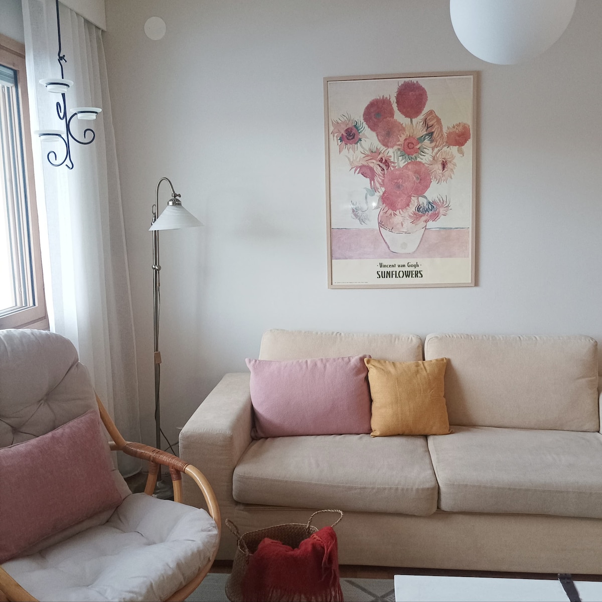 A light-filled living area features a beige sofa adorned with colorful cushions, including a soft pink and yellow pillow. A framed floral art piece is mounted on the wall, complementing a lamp and a cozy armchair nearby. Natural light pours in through a window with sheer curtains.