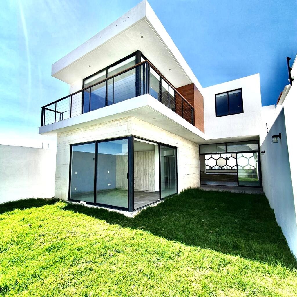 A contemporary two-story house features expansive glass walls that open to a green lawn. The upper level presents a balcony with a sleek railing, providing views over the spacious outdoor area. Natural light illuminates the interior through large windows.