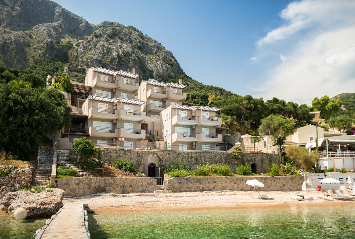 A cluster of modern villas is situated along the shoreline, featuring multiple balconies and large windows. The surrounding landscape includes lush greenery and mountainous terrain, providing a natural backdrop. A sandy beach and calm waters are visible in the foreground, enhancing the villa's serene setting.