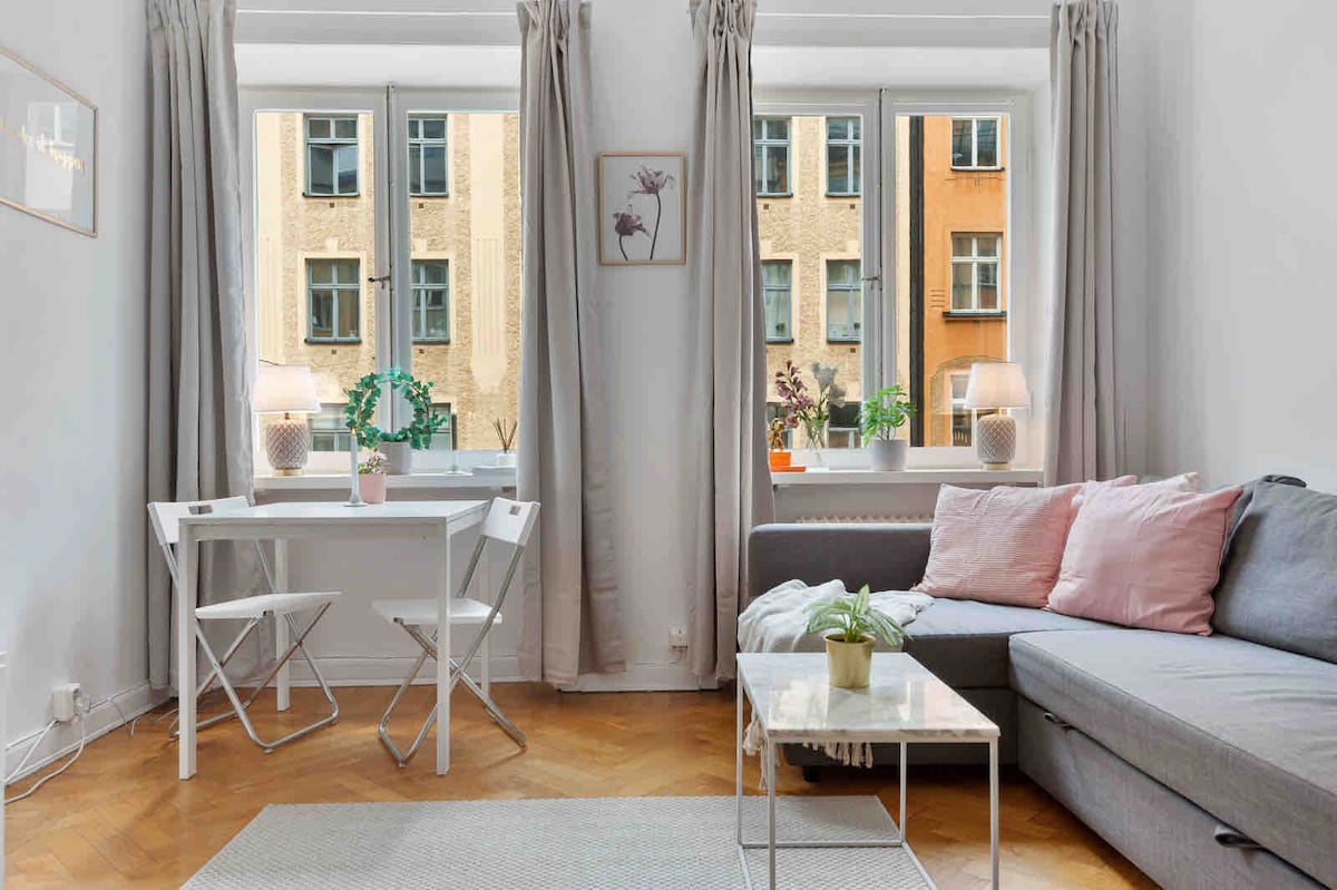 A welcoming living area features a gray sofa with soft pink pillows. A light-colored coffee table sits in front of the sofa. A compact white dining table with two folding chairs is positioned near the window, which is framed by light curtains. Natural light illuminates the space.