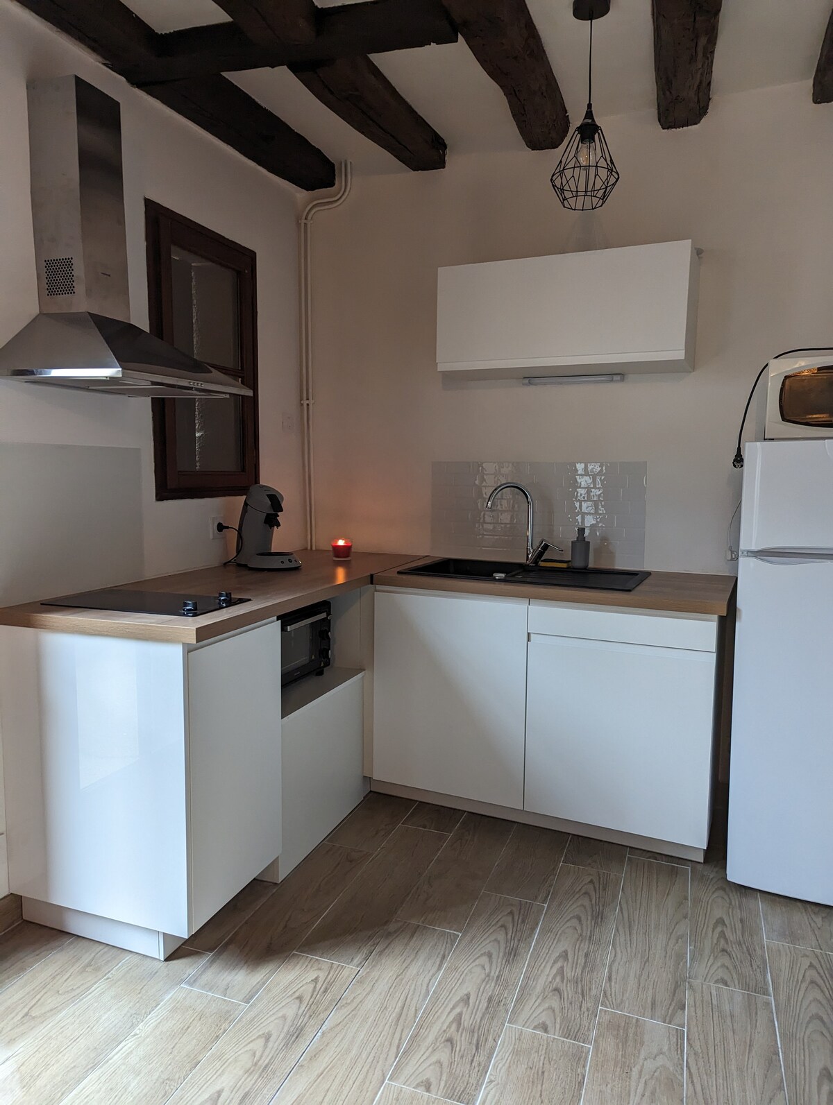 The kitchen features modern cabinetry in white, with a wooden countertop extending over the sink and stovetop. A stainless steel hood is positioned above the stovetop. Soft lighting is created by a candle, enhancing the clean and functional design of the space.