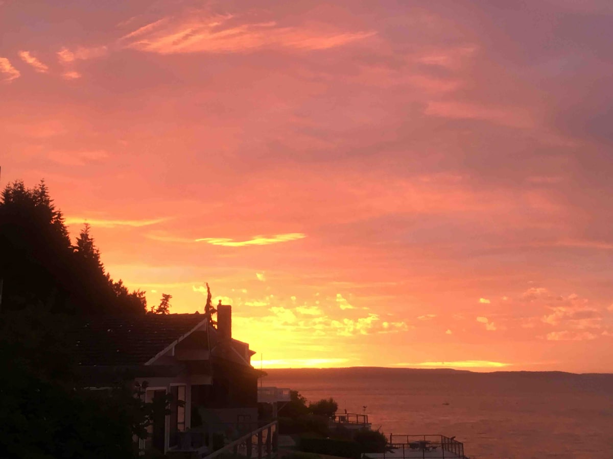 A warm sunset envelops the waterfront landscape, with vibrant hues of orange and pink filling the sky. The silhouette of the cottage is visible against the backdrop of the bay, with trees lining the horizon and water gently reflecting the colorful sky.