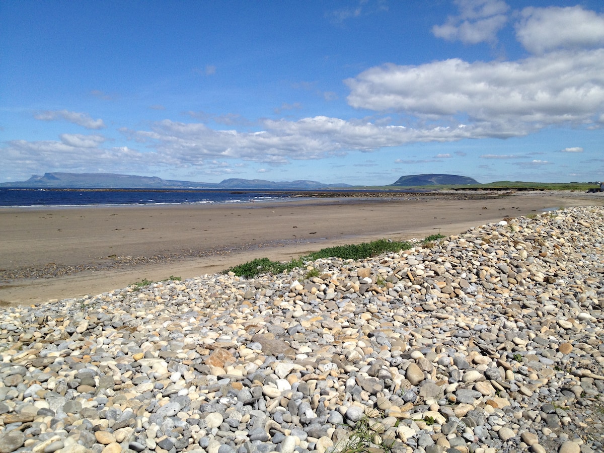 A sandy beach is framed by smooth, pebbled edges, leading to clear blue waters under a bright sky. Gentle waves can be seen lapping at the shore, with distant mountains providing a scenic backdrop.