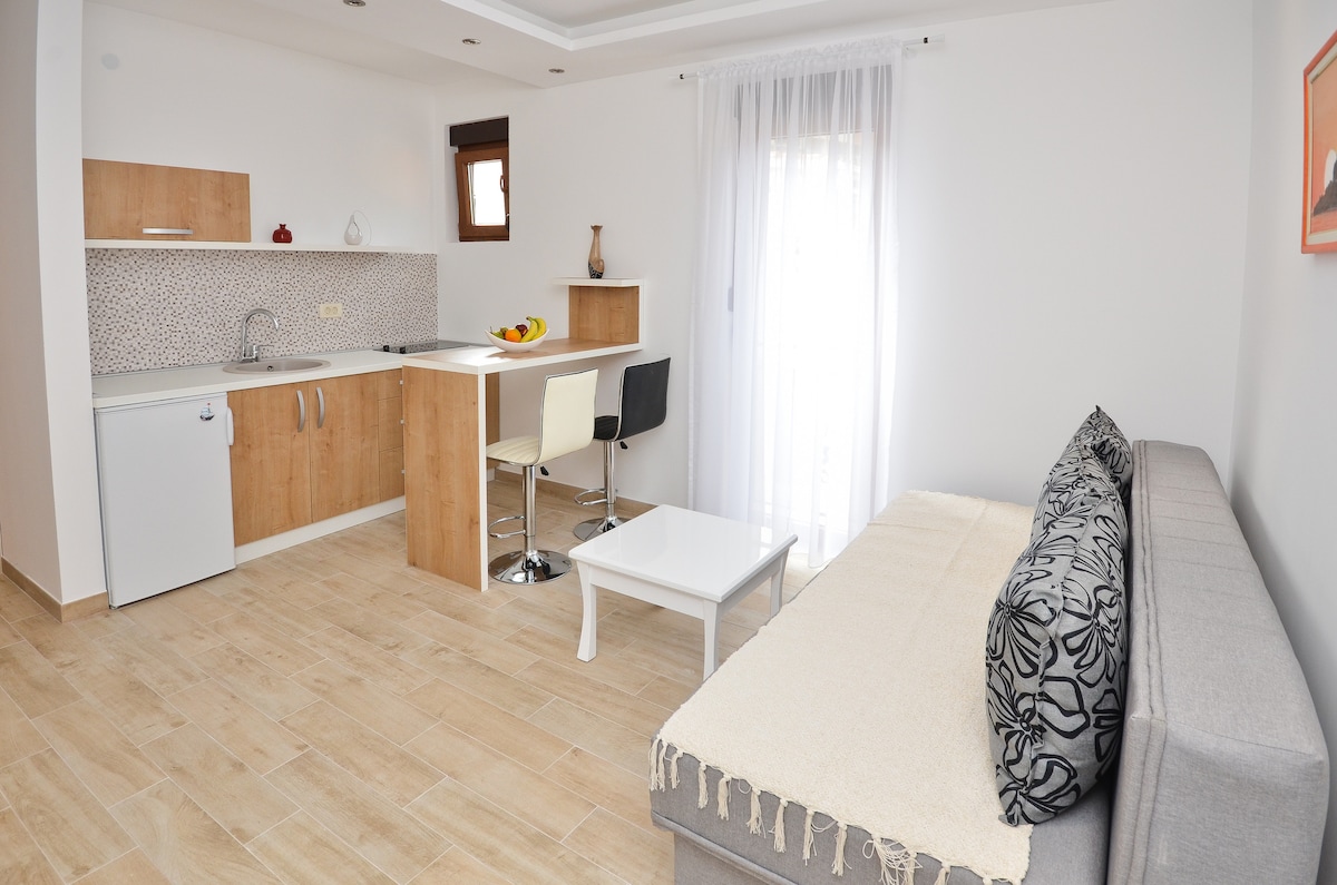 An inviting living area features a spacious sofa with decorative cushions, complemented by a light, neutral color palette. A modern kitchen area is visible in the background, with bar seating and a small dining table. Natural light filters in through a window with sheer curtains.