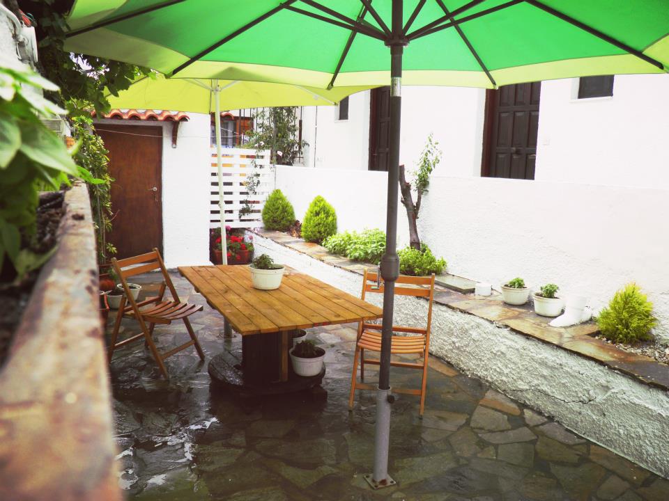 A shaded outdoor seating area is defined by a wooden table surrounded by four chairs. A large green umbrella provides shelter, while potted plants add greenery to the space. The stone flooring enhances the relaxed atmosphere of this tranquil garden setting.