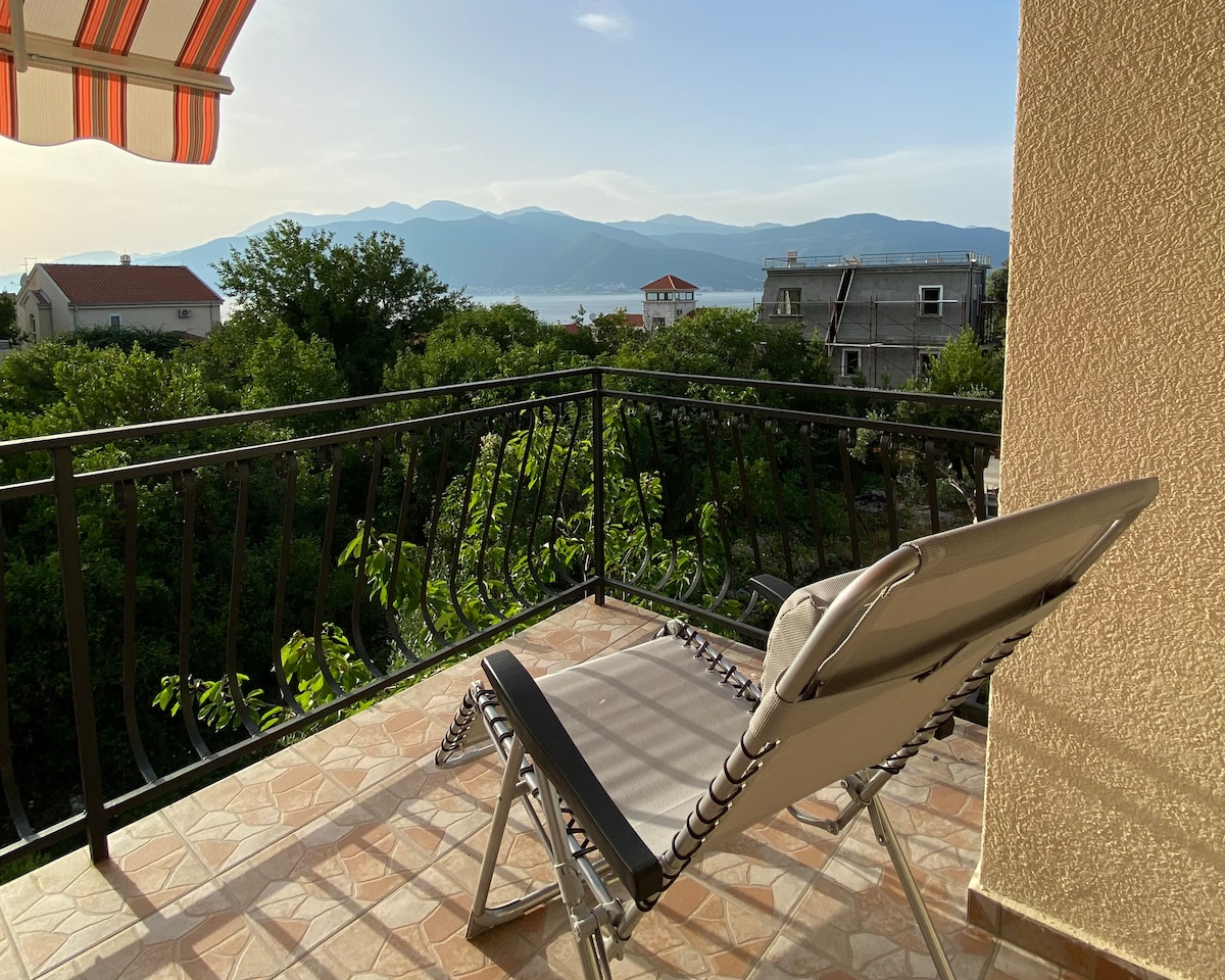 A balcony is featured with a folding lounge chair positioned for scenic views of the mountains and distant water. Lush greenery surrounds the space, enhancing the outdoor area that is adorned with patterned tile flooring. An awning provides shade from the sun.