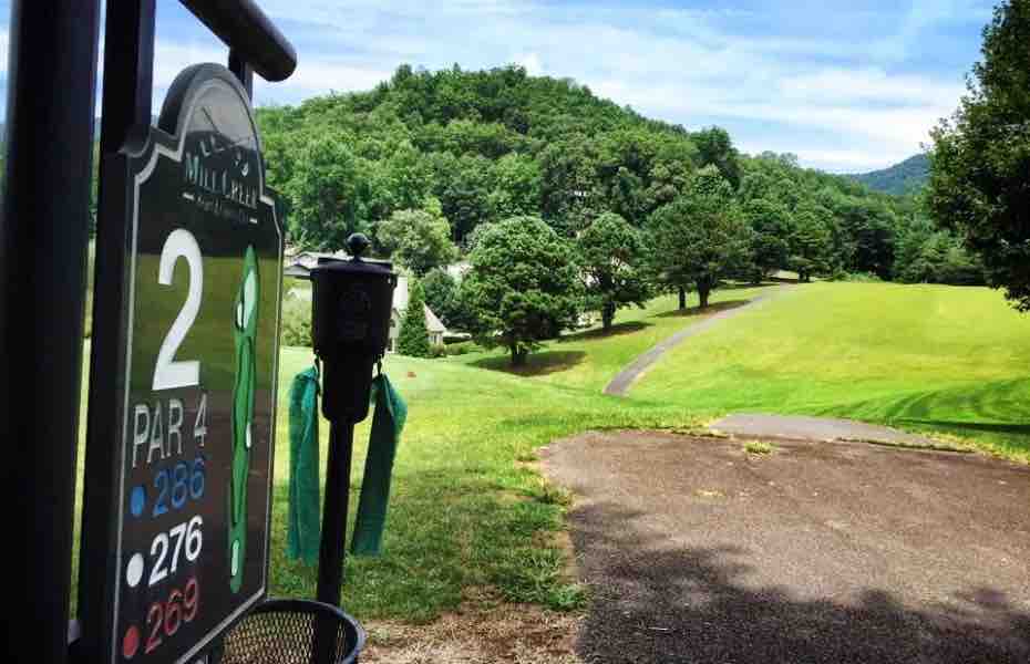 A golf course scene is depicted, featuring a numbered sign marking the second hole. The sign lists the hole as a par 4 with multiple tee options. A well-manicured fairway stretches into the distance, bordered by trees and rolling hills under a clear blue sky.