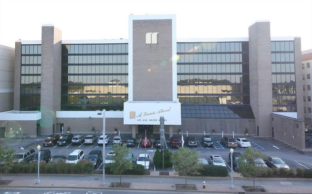 The exterior of Barclay Towers is presented, showcasing a modern multi-story building with large windows reflecting the surroundings. The entrance is marked with a welcoming sign, and a parking lot is visible in the foreground, featuring a mix of vehicles. Lush greenery can be seen at the base of the building.