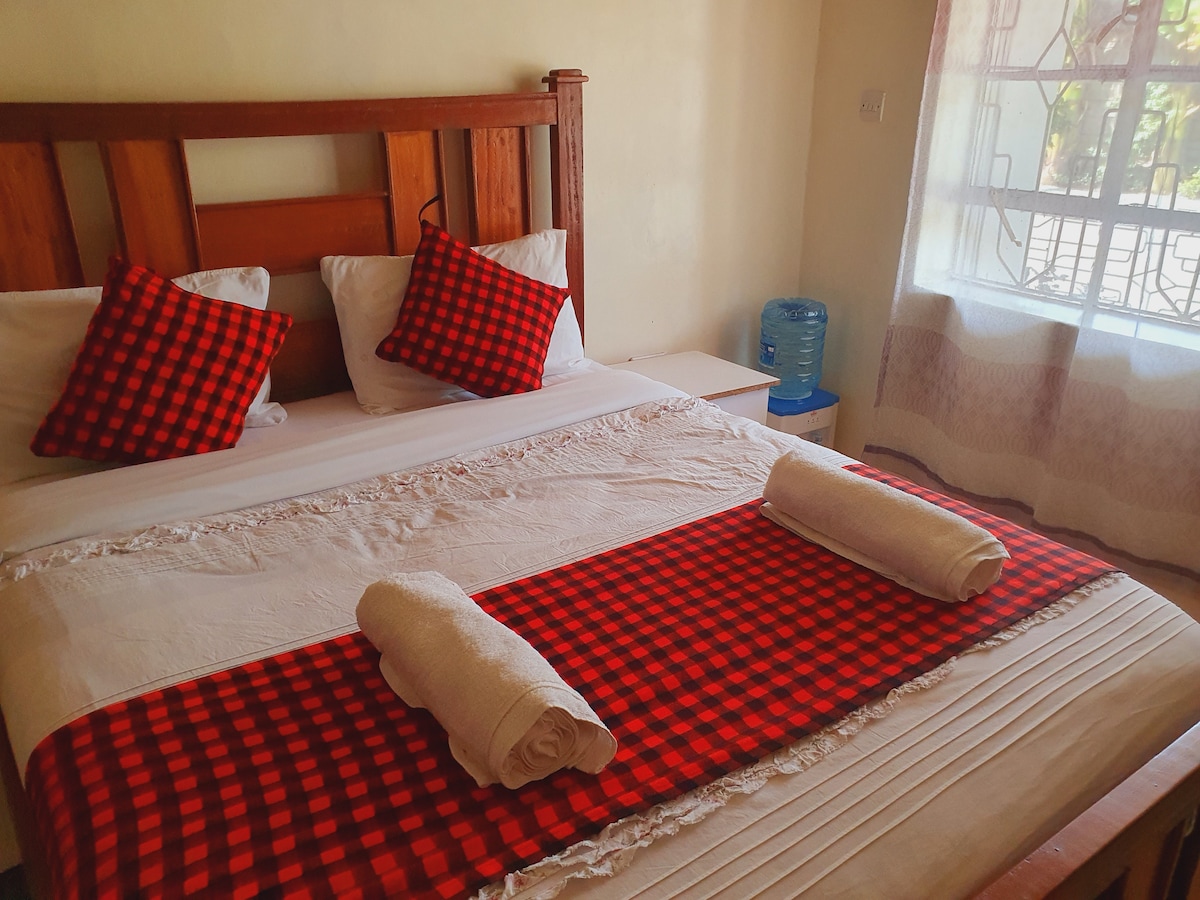 A well-arranged sleeping area is highlighted by a wooden headboard and red checkered accents on the bed. Two rolled towels are placed at the foot of the bed, and vibrant patterned pillows add a touch of colour. Natural light filters through a nearby window adorned with sheer curtains.