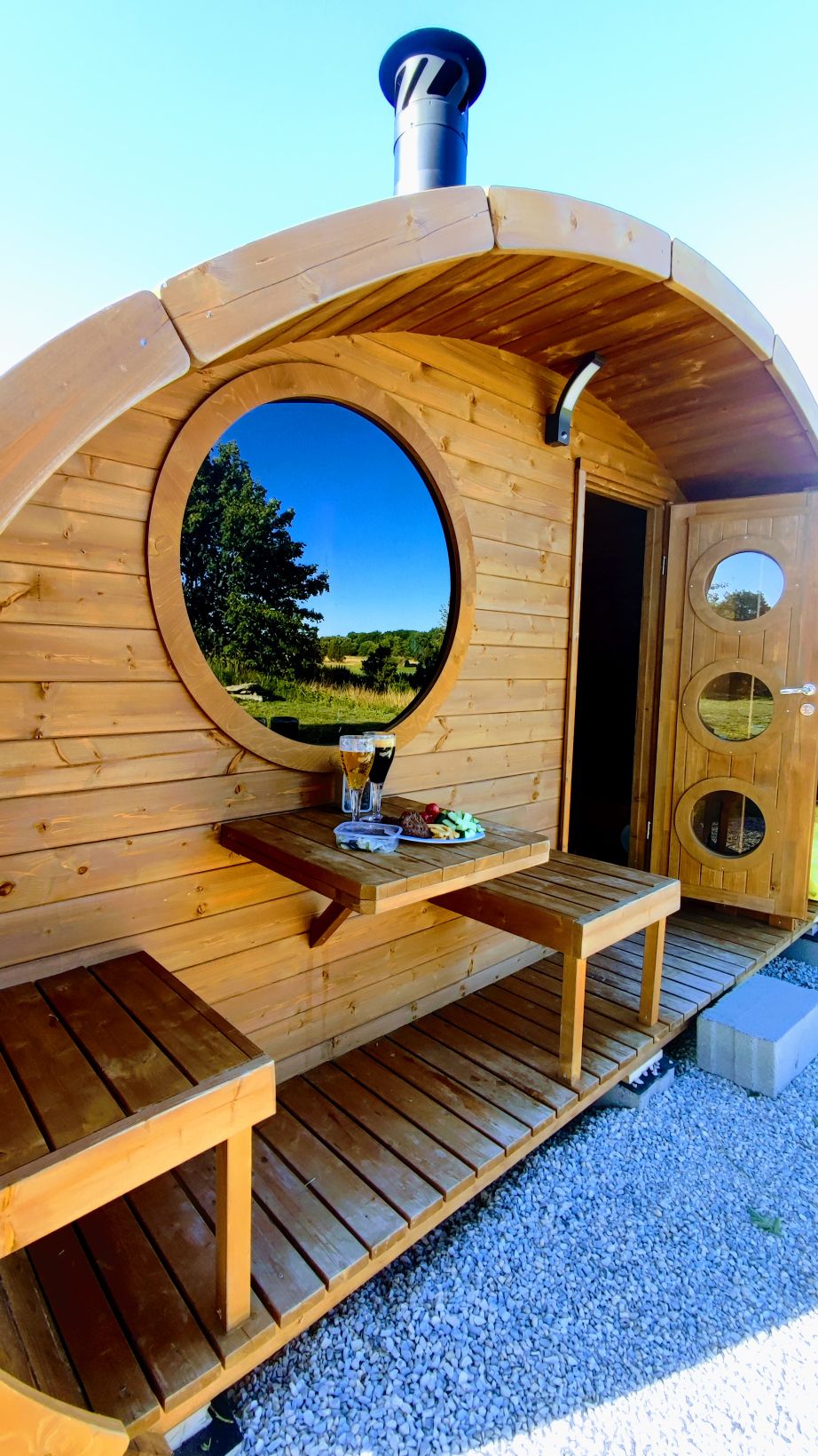A wooden sauna cabin features a rounded design with a large circular window providing views of the surrounding nature. Two wooden benches, positioned at the entrance, are complemented by a small table displaying refreshments. The door showcases three circular windows, enhancing the cabin's inviting aesthetic.