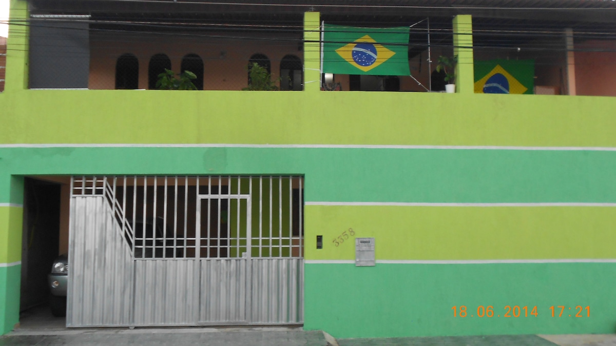 A vibrant green exterior of a multi-story accommodation is visible, featuring a secure, gated entrance. Brazilian flags are displayed on the upper level, and well-maintained plants can be seen through the arches of the balcony. Parking space for one vehicle is indicated.