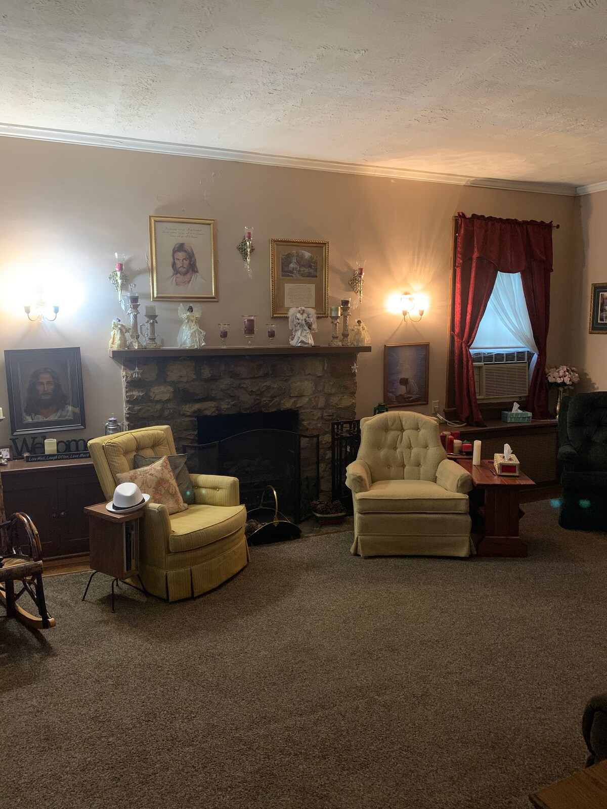 A cozy living room is presented, featuring a stone fireplace as a focal point. Two upholstered chairs in soft yellow are positioned nearby, alongside a wooden side table. Wall-mounted lights and framed artwork enhance the comfortable environment, while rich red curtains frame the windows.