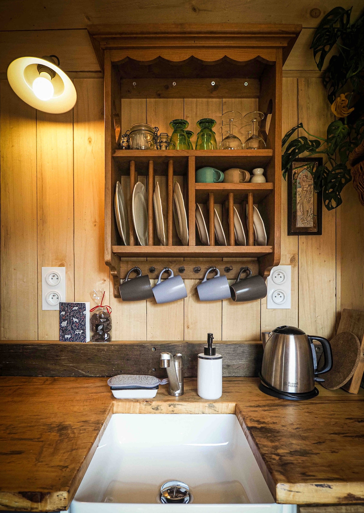 A wood-paneled kitchen area features a rustic sink beneath a shelf displaying neatly arranged plates and glassware. Hanging hooks hold several mugs, while a kettle sits on the counter beside a modern faucet. Soft lighting enhances the inviting atmosphere.