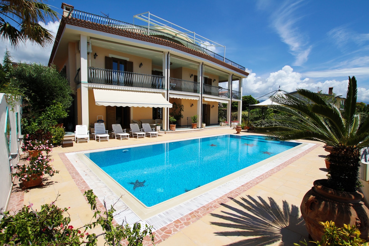 A spacious villa is presented, featuring a large outdoor swimming pool surrounded by lounge chairs. Lush greenery and potted plants border the pool area, while a covered terrace provides shade. The second floor balcony overlooks the serene setting under a clear blue sky.