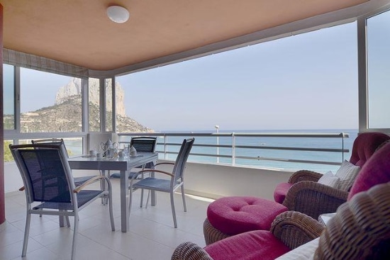 A spacious balcony offers a table with chairs, providing a setting for dining or relaxation. Comfortable lounge chairs are placed nearby for informal seating. The view showcases hills and the sea, visible through large glass windows that invite natural light.