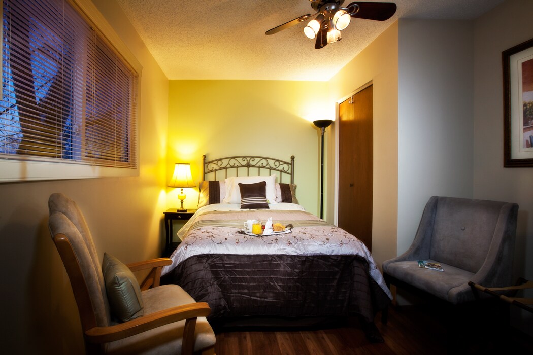 A cozy bedroom is highlighted by a spacious bed dressed in neutral linens. Two chairs are positioned nearby, and a bedside table supports a lamp. Warm light illuminates the room, emphasizing the soft colors of the walls and the dark wood flooring.