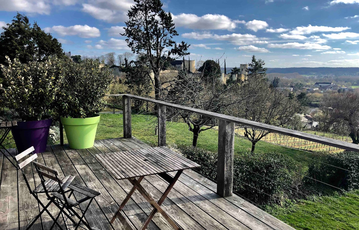 A wooden terrace is displayed, featuring a small folding table surrounded by two chairs. In the background, green and purple planters add color, while a panoramic view reveals the valley and distant castle. Lush greenery stretches beyond, enhancing the serene outdoor setting.